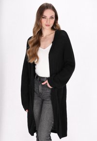 Schwarzer langer Cardigan mit strukturiertem Strick. Mit offenem Vorderteil, weiten Ärmeln und seitlichen Taschen, kombiniert mit einem weißen Oberteil und grauen Jeans.