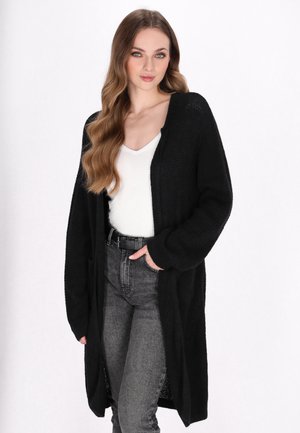 Schwarzer langer Cardigan mit strukturiertem Strick. Mit offenem Vorderteil, weiten Ärmeln und seitlichen Taschen, kombiniert mit einem weißen Oberteil und grauen Jeans.