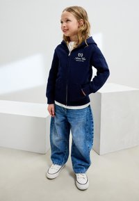 Sweat à capuche zippé bleu marine avec logo sur la poitrine, doté d'une capuche et de poches kangourou, accompagné d'un jean bleu ample et de baskets blanches.