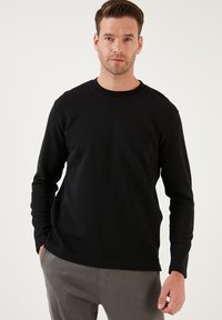 Fekete hosszú ujjú, kerek nyakú póló texturált mintával, könnyű anyagból készült. Szürke nadrággal párosítva. Simán illeszkedő, laza stílus.