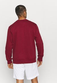 Homme portant un sweat-shirt bordeaux à manches longues et un short blanc, debout de dos contre un fond uni.