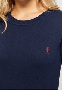 Námořnický modrý pletený svetr s kulatým výstřihem a červeným vyšívaným logem na levé hrudi. Měkká textura, přiléhavý střih.