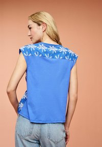 Blusenshirt aus blauem Baumwollstoff mit kurzen Ärmeln, verziert mit weißer Palmbaum-Stickerei über den Schultern. Abgerundeter Saum und lockere Passform.
