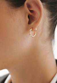 Pendiente de aro retorcido de oro exhibido en una oreja. El diseño presenta un acabado texturizado y un pequeño cierre, realzando su forma general.