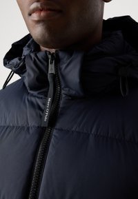Navy padded jacket with a high collar, featuring a zipper front and toggle details. Fabric has a smooth texture; logo tag is visible.