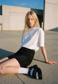 Jeune femme aux longs cheveux blonds portant un t-shirt blanc, une jupe noire, des chaussettes blanches et des sandales noires, assise sur du béton en plein soleil.