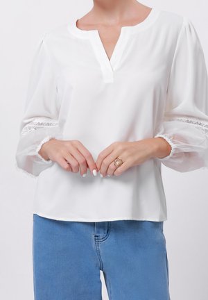 Kobieta ubrana w białą bluzkę z przezroczystymi koronkowymi rękawami i niebieskie dżinsy, z rękami splecionymi, na których widać złoty pierścionek z perłą.