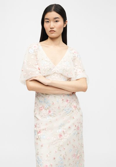 Γυναίκα με μακριά μαύρα μαλλιά, φορώντας ένα διάφανο φόρεμα με κεντήματα από λουλούδια και κοντά φουσκωτά μανίκια, στέκεται με σταυρωμένα τα χέρια μπροστά σε ένα λευκό φόντο.