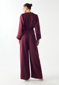 Burgundska široka kombinezona s dugim rukavima, remenom u struku, glatkom teksturom i detaljem s patentnim zatvaračem na leđima. Minimalistički dizajn, bez uzoraka.