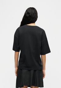 Γυναίκα με μακριά μαύρα μαλλιά, φορώντας χαλαρό μαύρο T-shirt και πλισέ μαύρη φούστα, γυρισμένη με την πλάτη προς το απλό ανοιχτό φόντο.