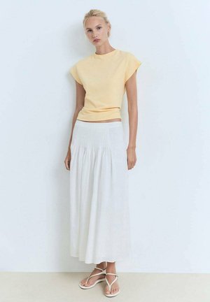 Top giallo a maniche corte abbinato a una gonna lunga e fluida bianca con pieghe. Sandali bianchi con cinturini completano l'outfit.