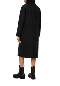 Schwarzer Wollmantel mit langen Ärmeln, taillierter Silhouette und seitlichen Taschen; kombiniert mit schwarzen Leder-Ankle-Boots mit klobigen Sohlen.