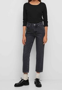 Zwarte langemouwen top gecombineerd met hoog-gewijde, enkel-lange grijze jeans en zwarte veterschoenen. Sokken zijn roomkleurig. Minimalistisch ontwerp.