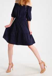 Schulterfreies navyblaues Kleid mit kurzen Puffärmeln, gerafftem Taillenteil und gestuftem Rock. Getragen mit nudefarbenen High Heels.