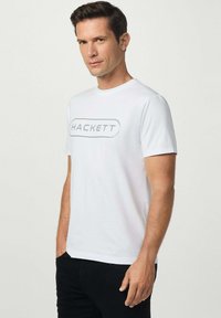 Camiseta de algodón blanco con un logo gris "HACKETT" en el centro. Diseño de manga corta y cuello redondo con una textura de tela suave.