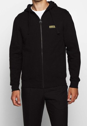 Mężczyzna ubrany w czarną bluzę z kapturem zapinaną na suwak, z sznurkami kaptura i małym żółtym logo „Barbour International” na piersi, na białej koszuli.