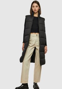 Gilet rembourré noir avec col haut et fermeture éclair, présentant un design long et texturé ainsi que des panneaux matelassés, porté sur un haut noir et un pantalon crème.