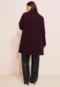 Un palton din lână burgundy, până la genunchi, cu guler, purtat peste pantaloni din piele neagră, având un design elegant și detalii minime.