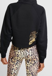 Person wearing a black cropped hoodie with gold Nike logos and leopard print leggings, shown from the back against a plain background.