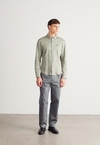 Svetlo zelena srajca z gumbi in ovratnikom, sive hlače in črni čevlji. Gladek tekstura z nežnim blagovnim znakom na srajci.