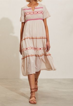 Nainen, jolla on yllään vaalea beige midi-mekko, jossa on vaaleanpunaisia ja punaisia brodeerattuja kuvioita, yhdistettynä ruskeisiin hihna-kenkiin, kävelee sisätiloissa.