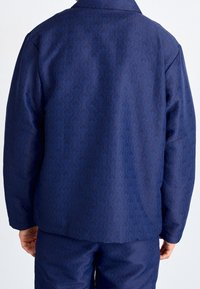 Mörkblå jacka med ett texturerat geometriskt mönster. Har en krage och långa ärmar, tillverkad av ett mjukt, lätt tyg.