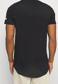 Czarna koszulka z krótkim rękawem wykonana z bawełny, z zakrzywionym dołem i białym napisem na lewym rękawie. Gładka tekstura.