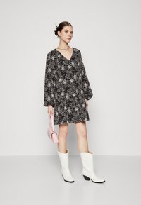 Robe noire à motifs floraux avec des manches longues, un décolleté en V et un ourlet ludique. Associée à des bottes de cowboy blanches et un petit sac à main rose avec des détails.