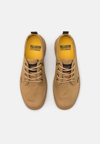 Gaišu brūnu audekla sporta apavu, ar gumijas pirkstu daļu, ar saistīšanas dizainu un dzeltenu iekšzoli, uz kuras ir melna "PALLADIUM" zīmola nosaukums.