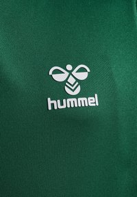 Groene sportstof met ribbelstructuur, met een witte Hummel-logo en vlinderembleem, centraal geplaatst.