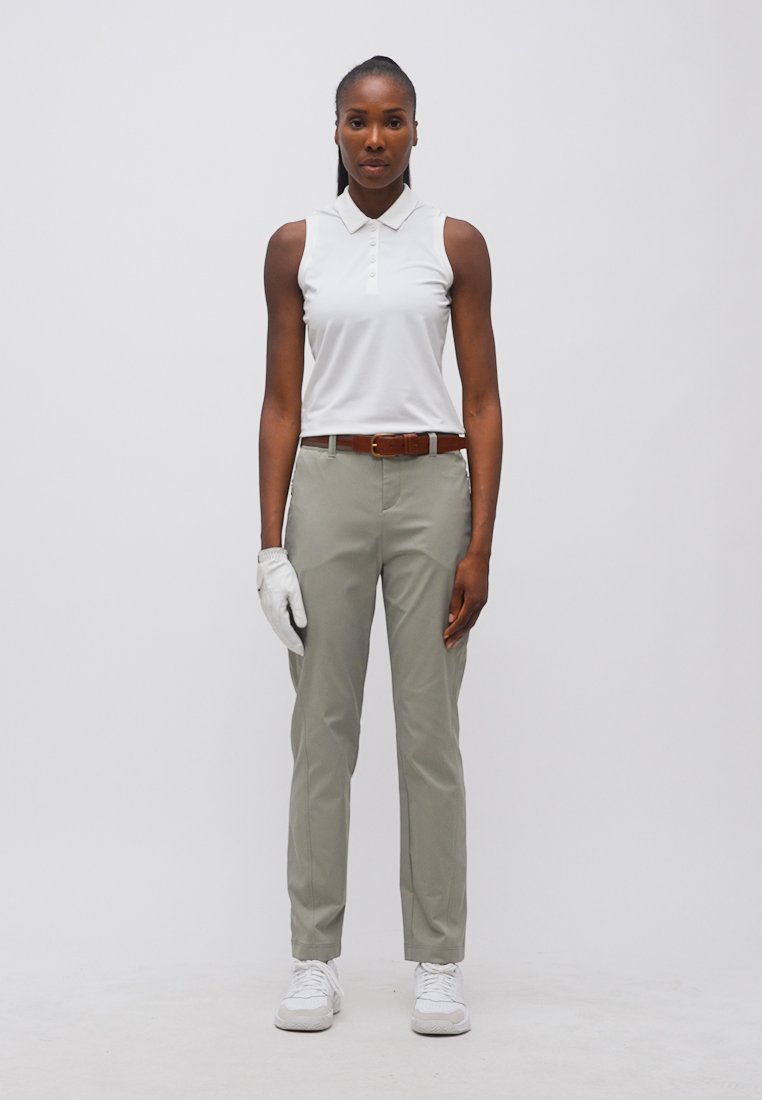 Femme en polo blanc sans manches, pantalon gris clair, ceinture marron, baskets blanches et gant de golf blanc à la main gauche, debout devant un fond uni.