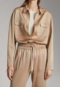 Chemise beige à boutons avec deux poches poitrine, associée à un pantalon à taille élastique avec cordon de serrage assorti. Texture lisse, coupe décontractée.