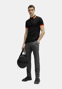 Polo nero con dettagli arancioni nel colletto, abbinato a pantaloni grigi. Modello che tiene una borsa rotonda nera, indossando scarpe da ginnastica nere.