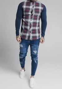 Red and blue checkered long-sleeve shirt with a buttoned front, paired with distressed blue jeans and white sneakers.