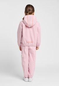 Roze jack met elastische manchetten en capuchon, voorzien van oranje accentstiksels. Gecombineerd met bijpassende roze broek en witte schoenen.