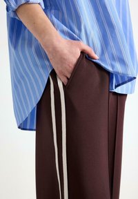 Camicia a righe blu con maniche arrotolate sopra pantaloni marroni con strisce laterali e una mano in tasca. Focus sulle trame dei tessuti.