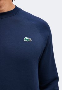 Chemise bleu marine à manches longues avec encolure ronde, montrant un petit logo crocodile vert sur la poitrine gauche.