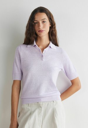 Kvinde med langt bølget hår iført en lyse lilla kortærmet trøje med krave og lyse beige bukser, stående mod en ensfarvet hvid væg.