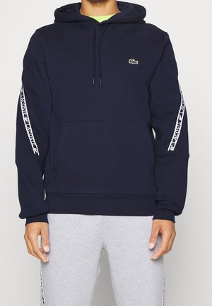 Muž v tmavě modré mikině s kapucí s bílou páskou s logem Lacoste na rukávech a malým zeleným krokodýlím emblémem na hrudi, doplněný světle šedými kalhotami.