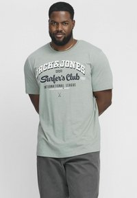 Gaiši zaļa īsām piedurknēm T-krekls ar baltiem un tumši zilajiem "Jack & Jones Surfer's Club" grafiskajiem elementiem. Mīksta tekstūra, brīva piegriezums. Pelēkas apakšdaļas komplekts.