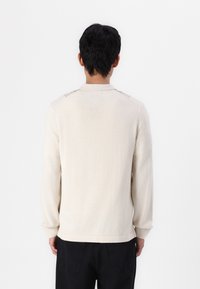 Cream-colored sweater made from soft fabric, featuring long sleeves and a subtle ribbed pattern along the shoulders and collar.