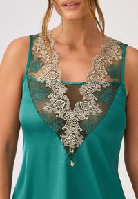 Teal satynowa bluzka z głębokim dekoltem w kształcie V, czarną koronką z kwiatowym wzorem i złotym akcentem na środku. Złożone detale koronkowe podkreślają fakturę.