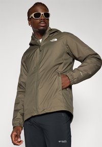 Veste légère vert olive avec capuche et poignets élastiques, dotée d'une fermeture éclair à l'avant et d'un détail de logo sur la poitrine. Associée à un pantalon sombre.
