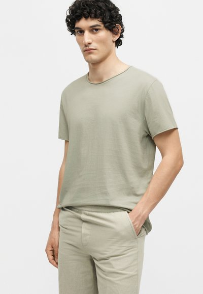 Άνδρας με κατσαρά μαλλιά που φοράει ένα ανοιχτό μπεζ κοντομάνικο μπλουζάκι και τσουλώσεις που ταιριάζουν, στέκεται με το ένα χέρι στην τσέπη σε λευκό φόντο.