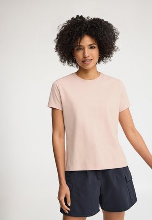 Kobieta z kręconymi włosami w jasnoróżowej koszulce z krótkim rękawem i ciemnoniebieskich krótkich spodenkach typu cargo, uśmiechająca się na jednolitym tle.