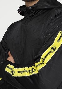 Svart nylonjacka med huva, med en texturerad yta, gula accentstripes med logotyp och dragsko-stängningar vid halsringningen.