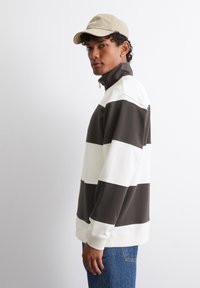 Barna és fehér csíkos pulóver magas nyakkal és cipzárral, bézs sapkával párosítva. Bordás mandzsettákkal és laza szabással rendelkezik.