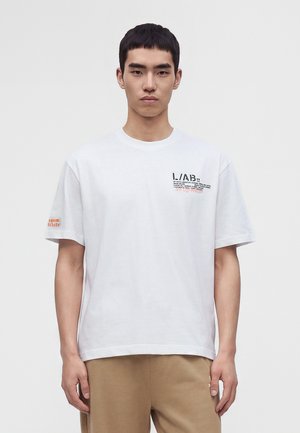 Mann trägt weißes kurzärmeliges T-Shirt mit schwarzem und rotem Text auf der Brust und kleinem orangefarbenem Text am linken Ärmel, kombiniert mit beigen Hosen.