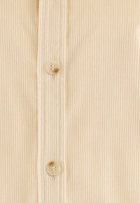 Svetle béžová košile z žebrované látky s knoflíkovou légou a světlými knoflíky, viditelným šitím podél okrajů.
