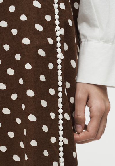 Material maro cu buline albe neregulate și o margine verticală cu pompoane rotunde albe, lângă o mână relaxată care poartă o cămașă albă cu manșete.